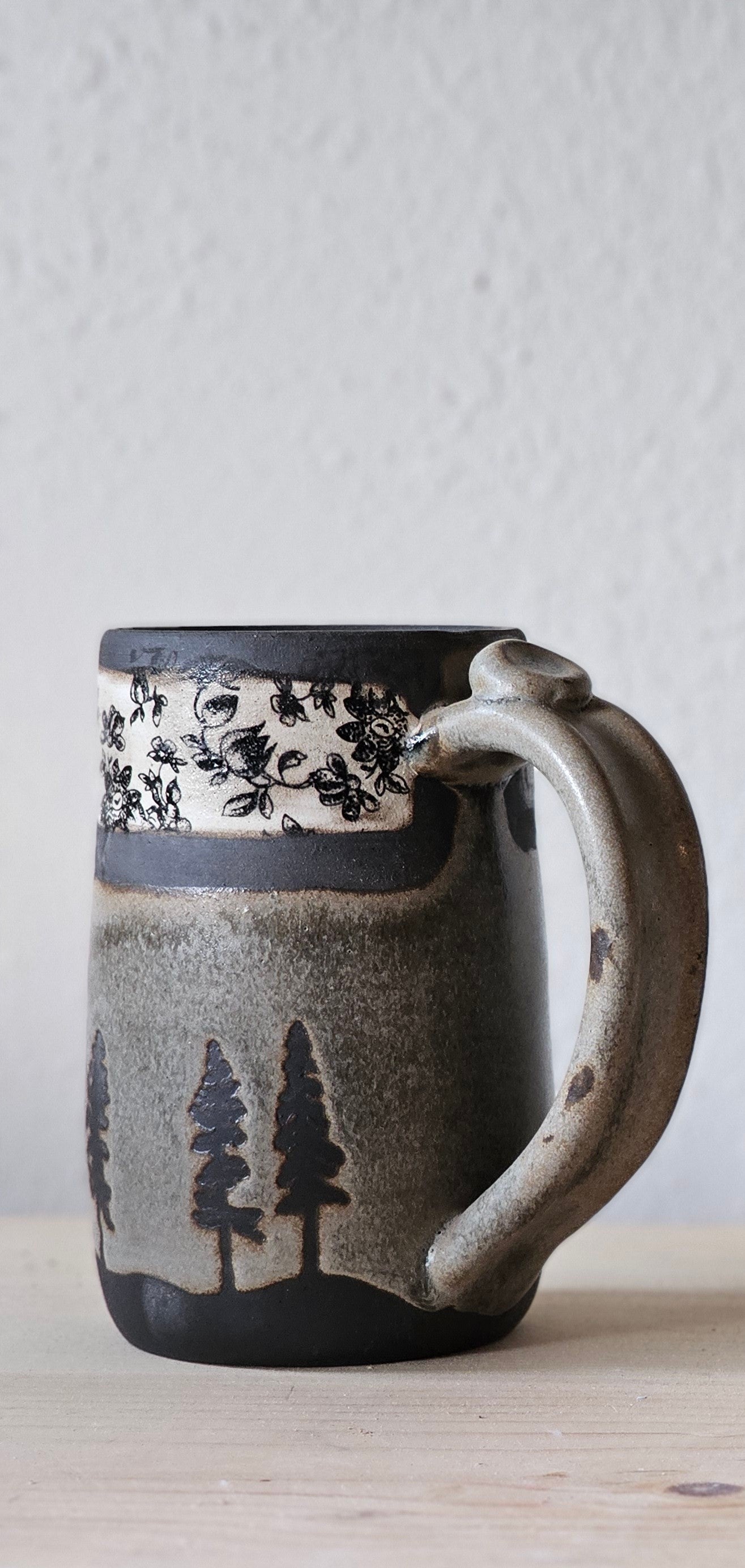 Woodland Heirloom ceramic mug in a taller profile, with earthy gray-green glaze, black evergreen trees , healthy 3 or 4 finger handle with thumb tab, and vintage inspired band.  
