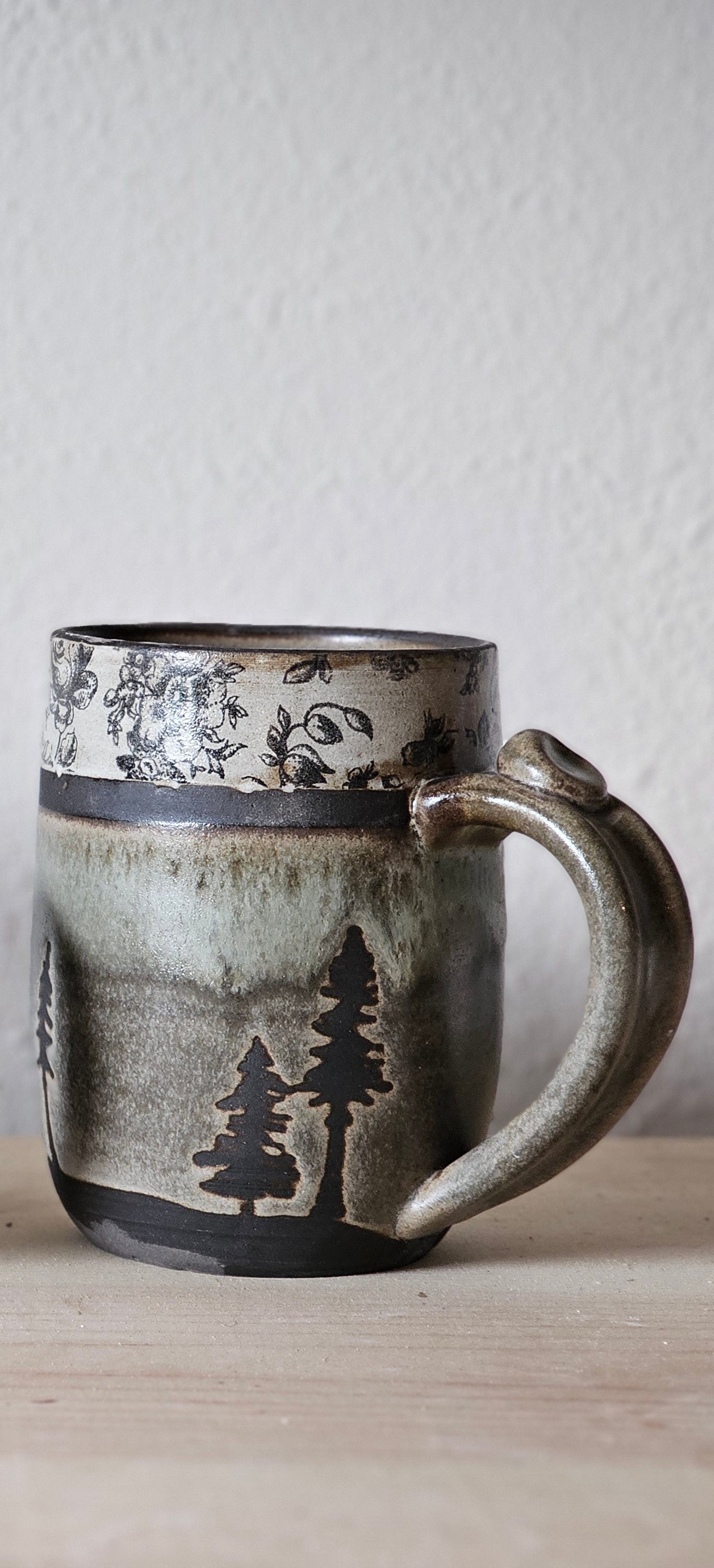 Woodland Heirloom ceramic mug with earthy gray-green glaze, black evergreen trees , healthy 3 finger handle with thumb tab, and vintage inspired band.  