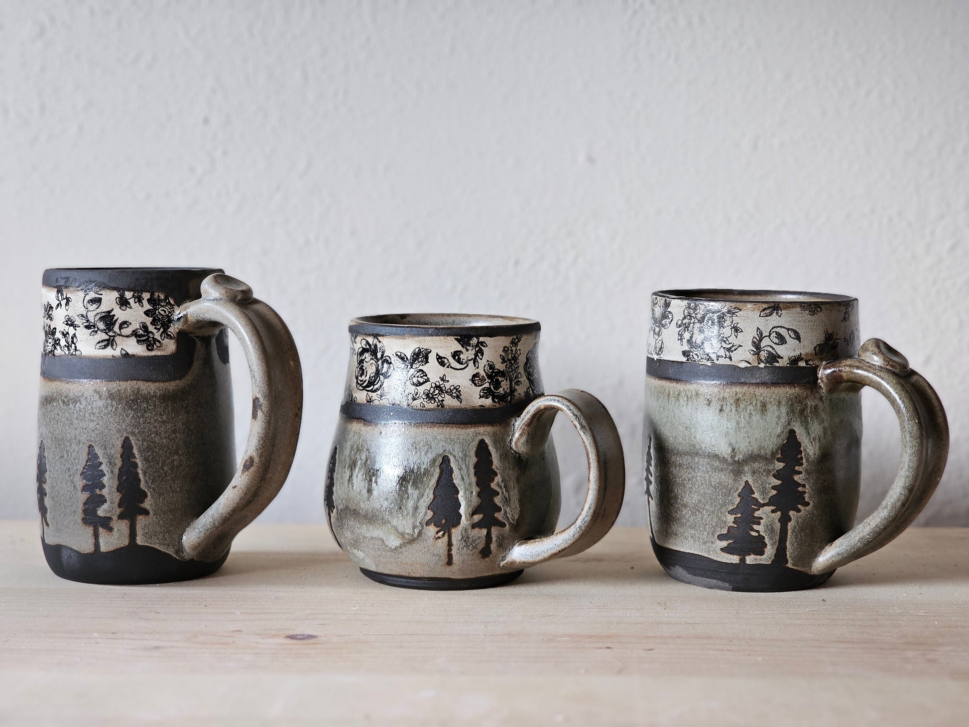 three handmade Woodland Heirloom ceramic mugs with earthy gray-green glazes, black evergreen trees and vintage inspired black floral bands.